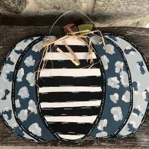 Puede incluir: Un colgador de puerta de calabaza de madera decorado con rayas blancas y negras y estampado de leopardo azul y blanco. La calabaza tiene un lazo de paja y un colgador de alambre.