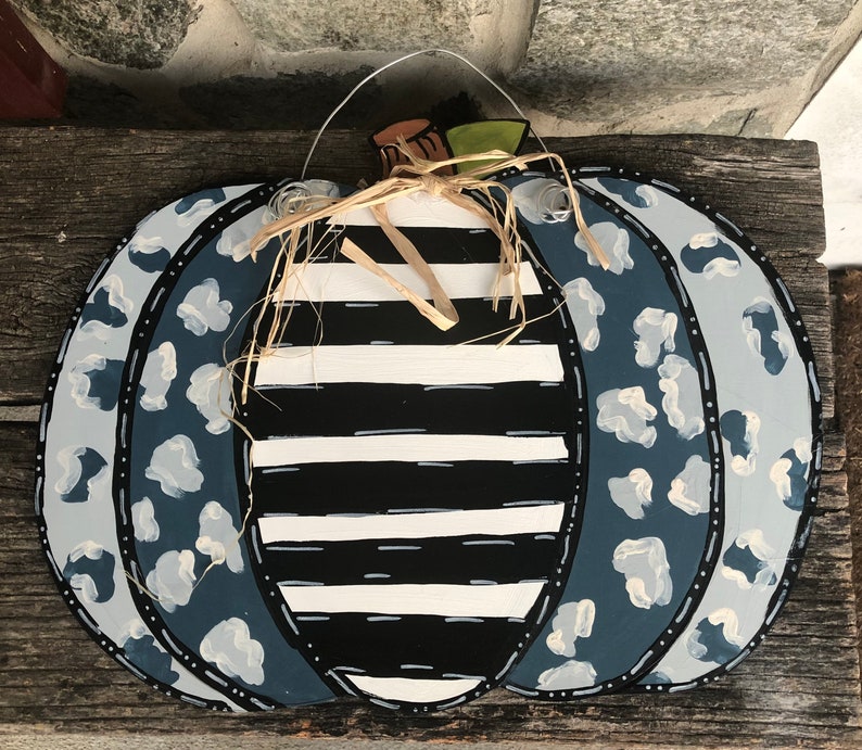 Puede incluir: Un colgador de puerta de calabaza de madera decorado con rayas blancas y negras y estampado de leopardo azul y blanco. La calabaza tiene un lazo de paja y un colgador de alambre.