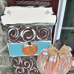 May include: A wooden coffee cup door hanger with a brown and white swirl design, a blue stripe, and a small orange pumpkin. A larger orange, blue, and brown pumpkin with a burlap bow is attached to the side.
