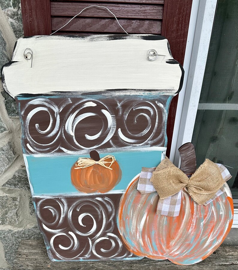 May include: A wooden coffee cup door hanger with a brown and white swirl design, a blue stripe, and a small orange pumpkin. A larger orange, blue, and brown pumpkin with a burlap bow is attached to the side.