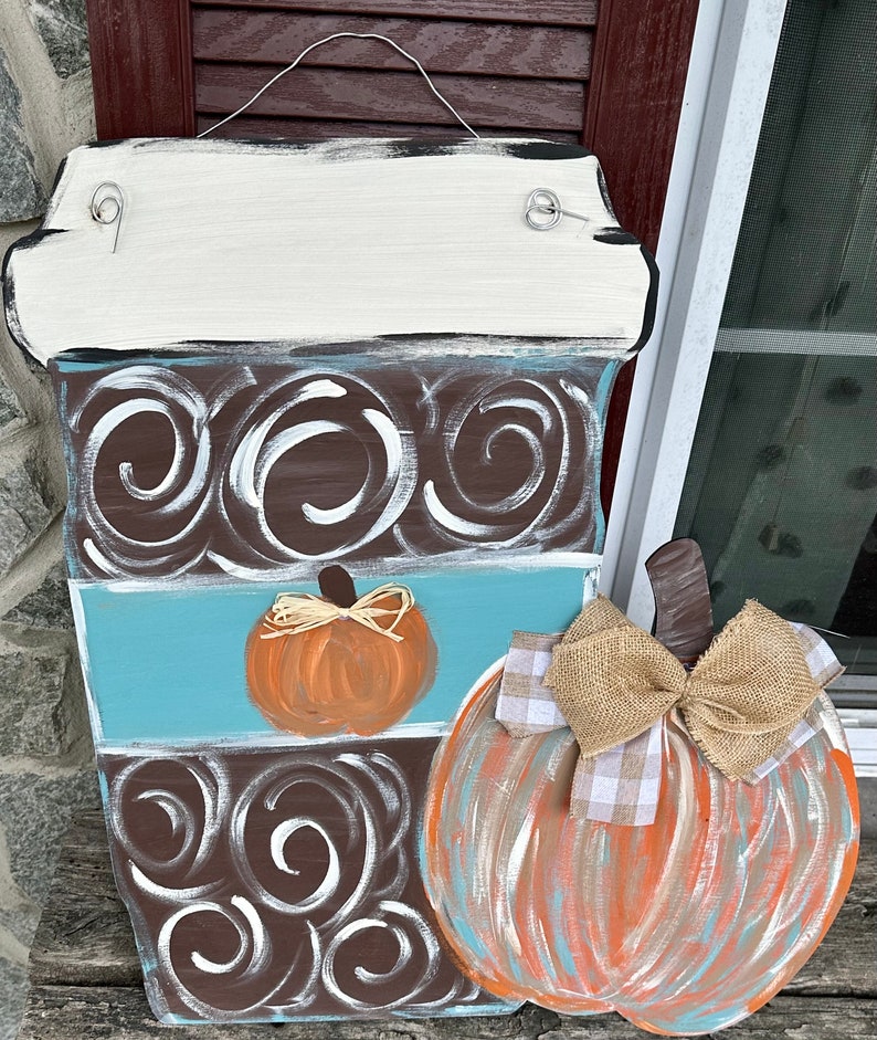 May include: A wooden coffee cup door hanger with a brown and white swirl design, a blue stripe, and a small orange pumpkin. A larger pumpkin with a burlap bow is attached to the side of the coffee cup.