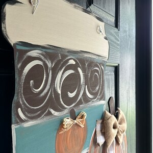 May include: A brown and blue painted wooden door hanger shaped like a coffee cup with a white lid. The cup has a swirl pattern and is decorated with two orange pumpkins with burlap bows.