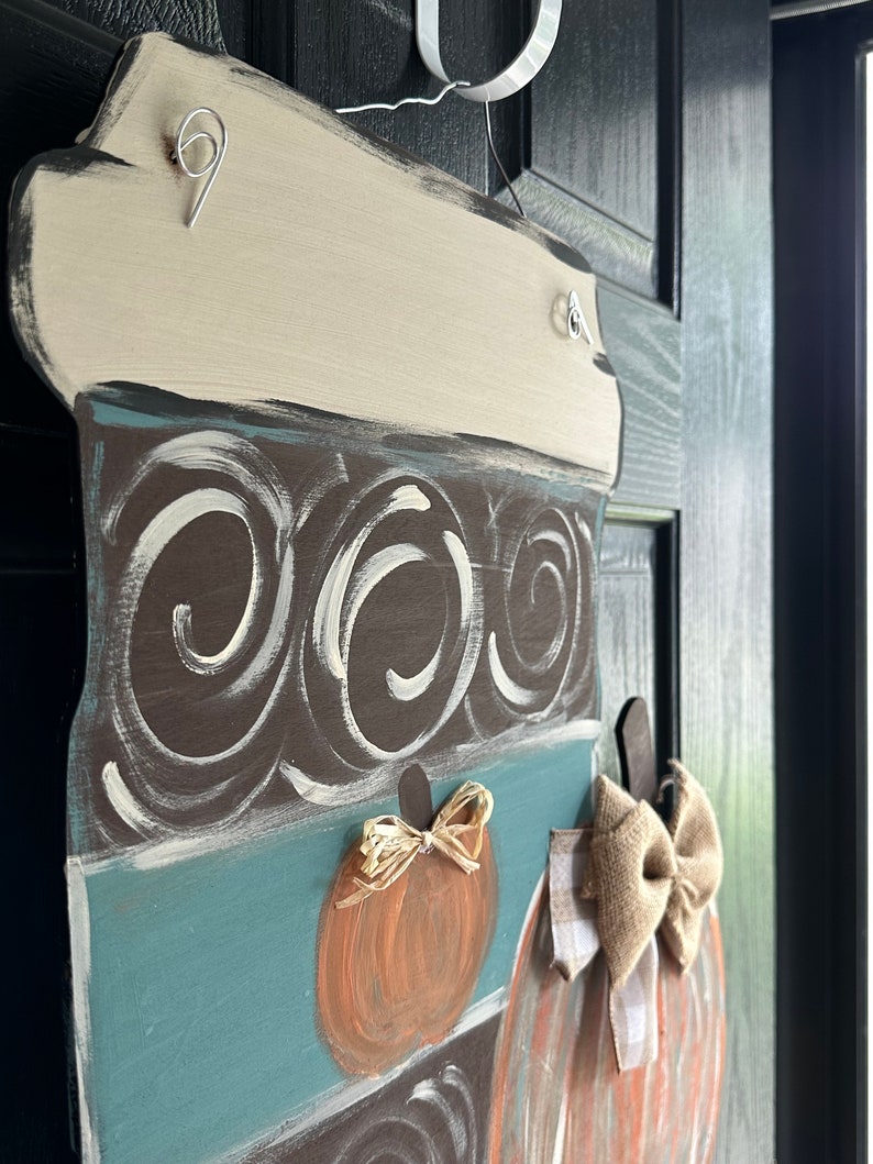 May include: A brown and blue painted wooden door hanger shaped like a coffee cup with a white lid. The cup has a swirl pattern and is decorated with two orange pumpkins with burlap bows.