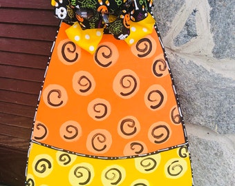 May include: A Halloween-themed candy corn door hanger with a yellow and orange swirl pattern. The door hanger is decorated with a black and white ribbon with a spooky Halloween print.