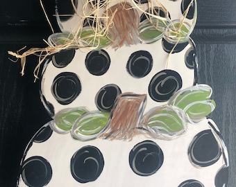 May include: A white, wooden pumpkin door hanger with black polka dots and green leaves. The pumpkin is stacked on top of two smaller pumpkins, and the top pumpkin has a brown stem and a bow made of natural twine.