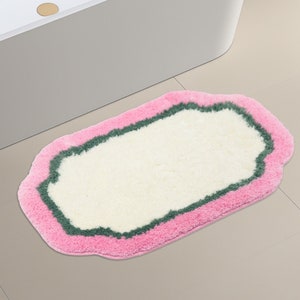 May include: A white bath mat with a pink and green border. The mat has a scalloped edge and is made of soft, fluffy material.