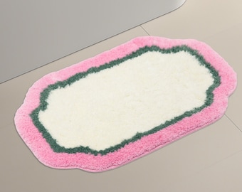 Puede incluir: Una alfombra de baño blanca con un borde rosa y verde. La alfombra tiene un borde festoneado y está hecha de un material suave y esponjoso.