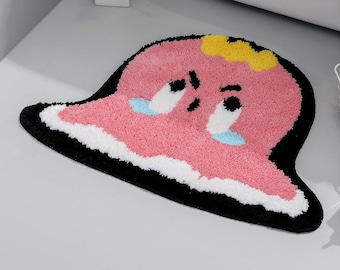 May include: Pink bath mat with a cartoon character design. The character has a yellow crown, big eyes, and a sad expression. The mat has a black border and white accents.