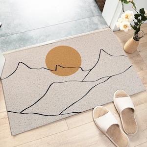 May include: A beige doormat with a minimalist design featuring a black outline of mountains and a yellow sun. The doormat is made of a textured material and has a subtle speckled pattern.
