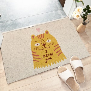 May include: A beige doormat with a yellow cat illustration. The cat has red stripes and says "I MEOW you".