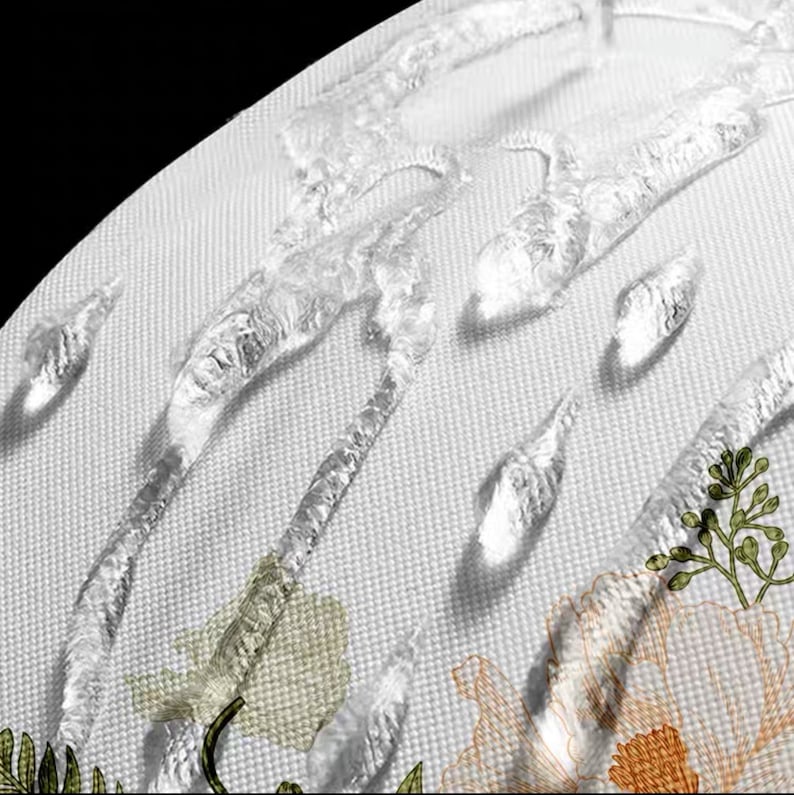 K&ouml;nnte beinhalten: Nahaufnahme eines wei&szlig;en Stoffes mit Wassertropfen und floraler Stickerei. Das Design zeigt eine hellbeige Blume mit orangefarbenen und gr&uuml;nen Akzenten und einen gr&uuml;nen Stiel. Die Wassertropfen sind klar und gl&auml;nzend.