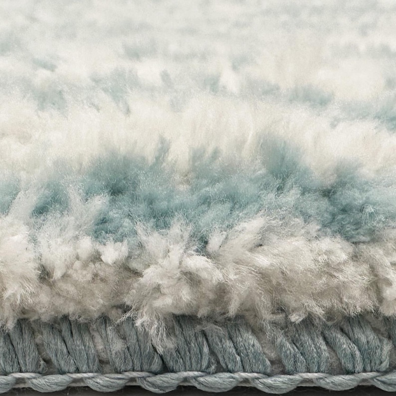 May include: Close-up of a soft, textured rug with a layered design. The rug features bands of white, light blue, and grey, creating a subtle, calming aesthetic. The bottom edge has a braided border in a matching blue-grey colour.