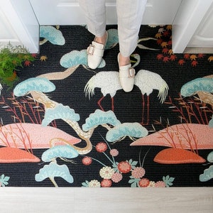 Puede incluir: Felpudo negro y blanco con un diseño de inspiración japonesa que presenta grullas, pinos y flores. El felpudo tiene una superficie texturizada.