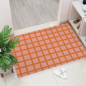 Puede incluir: Una alfombra rectangular con un patrón de cuadrícula en tonos naranja y rosa. La alfombra está colocada en una entrada, con zapatillas blancas cerca. Una planta en maceta es visible en el lado izquierdo.