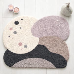 May include: A multi-colored bath mat with an abstract design. The mat features cream, lavender, gray, and black sections with dotted accents. The mat is made of a soft, plush material.