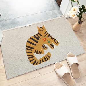 May include: A white doormat with a brown and black striped cat illustration holding a red flower. The cat is smiling and has a white background.