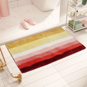 May include: A rectangular bath mat with horizontal stripes in shades of yellow, white, pink, and red. The mat is made of a soft, plush material and is placed on a white tiled floor. The bathroom setting includes a shower head, slippers, and a shelf with towels.