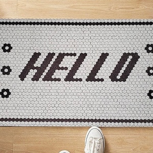 Puede incluir: Una alfombra blanca para la puerta con un borde negro y la palabra "HELLO" en letras negras. La alfombra está diseñada para parecer un mosaico de pequeños hexágonos blancos.