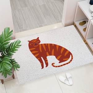 May include: A rectangular doormat with a white speckled background and a stylized orange and brown tiger illustration. The tiger is in a seated position with its tail curled. White slippers are placed near the mat.
