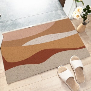 Könnte beinhalten: Eine beige, braune und rote abstrakte Türmatte mit einem wellenförmigen Design. Die Fußmatte liegt auf einem hellen Holzboden.