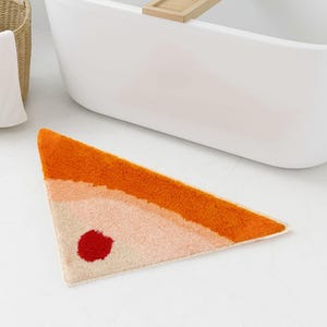 Puede incluir: Alfombra de baño triangular con un diseño degradado en tonos naranja, melocotón y beige, con un círculo rojo. La alfombra está hecha de un material suave y texturizado, colocada sobre un suelo de baldosas blancas.