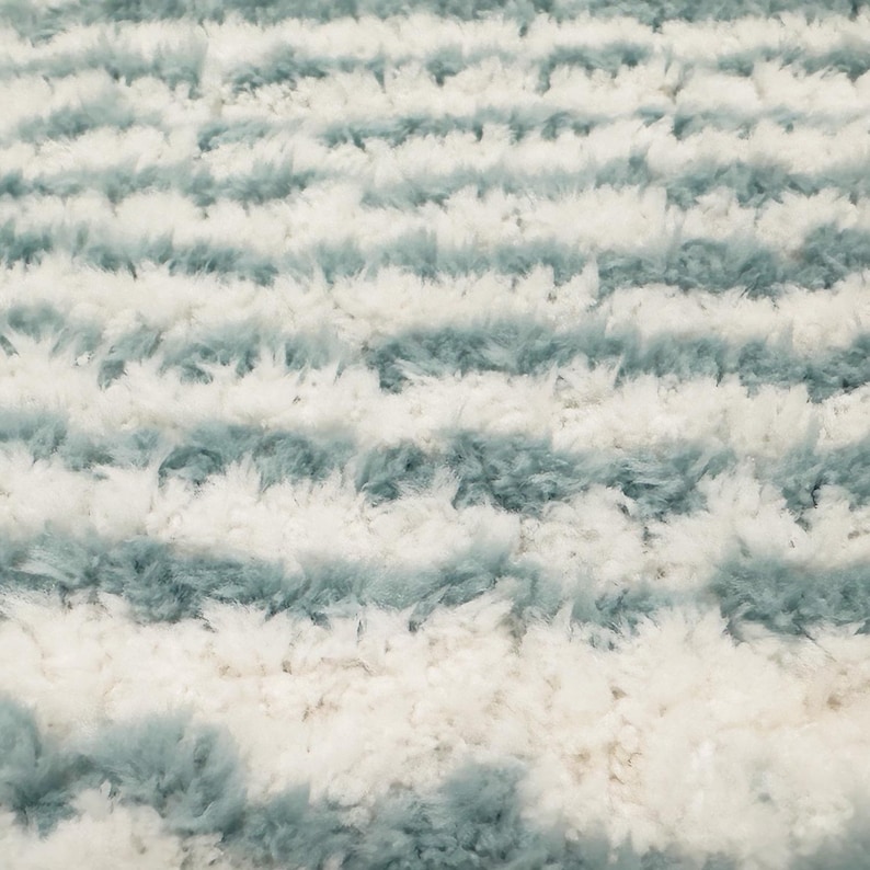 May include: A close-up view of a soft rug with a striped pattern. The rug has alternating bands of white and pale blue-green, creating a plush, inviting surface. The fibres appear fluffy and dense, suggesting a comfortable texture.