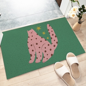 May include: A green doormat with a pink and brown spotted cat design. The cat is facing to the right and has a long tail. There are small yellow stars scattered around the cat.