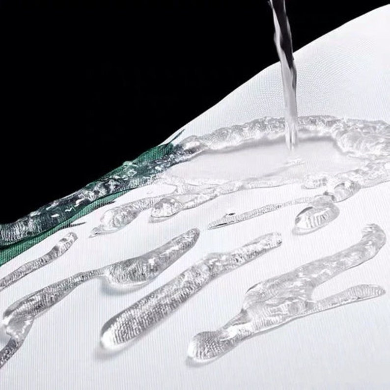 K&ouml;nnte beinhalten: Nahaufnahme von Wassertropfen auf einem wei&szlig;en Stoff. Wasser wird auf den Stoff gegossen, was die Wasserbest&auml;ndigkeit demonstriert. Der Stoff hat ein gr&uuml;n-wei&szlig;es Muster am Rand.