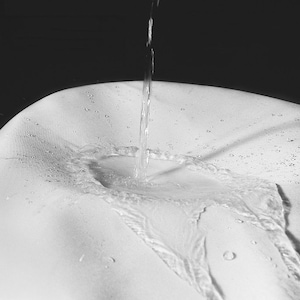 K&ouml;nnte beinhalten: Ein Graustufenbild von Wasser, das auf eine wei&szlig;e Oberfl&auml;che gegossen wird. Der Wasserstrahl erzeugt einen dynamischen visuellen Effekt, mit sichtbaren Tropfen und Wellen. Der Kontrast zwischen Wasser und Oberfl&auml;che hebt Textur und Form hervor.