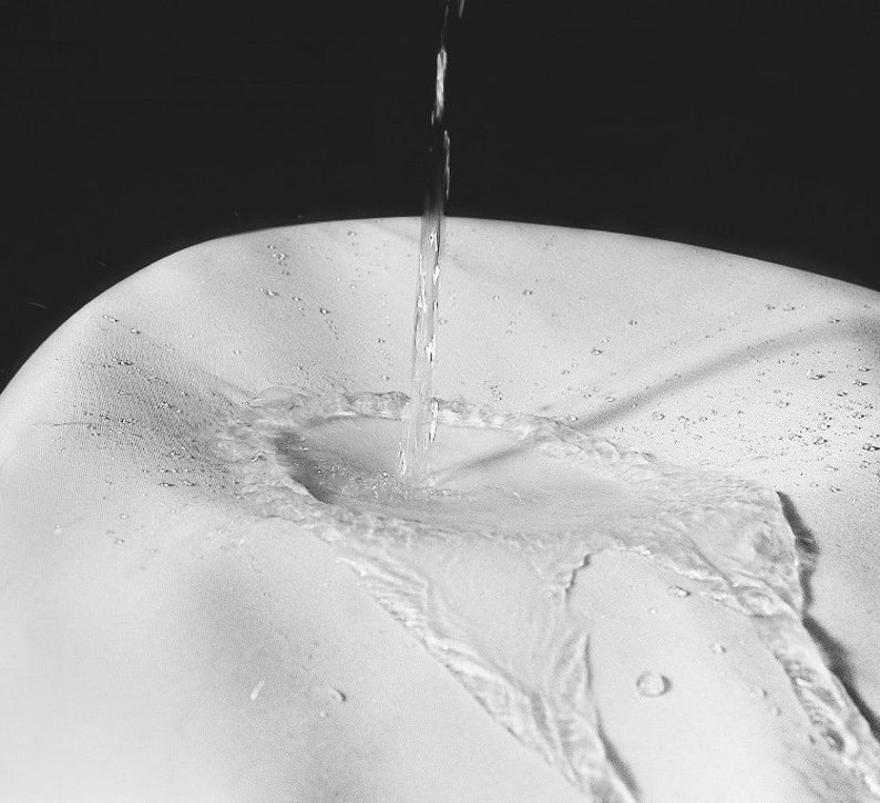 K&ouml;nnte beinhalten: Ein Graustufenbild von Wasser, das auf eine wei&szlig;e Oberfl&auml;che gegossen wird. Der Wasserstrahl erzeugt einen dynamischen visuellen Effekt, mit sichtbaren Tropfen und Wellen. Der Kontrast zwischen Wasser und Oberfl&auml;che hebt Textur und Form hervor.