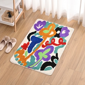 Könnte beinhalten: Eine weiße Badematte mit einem farbenfrohen abstrakten Blumendesign. Das Design zeigt orange, blaue, grüne, violette, rote und schwarze Formen.