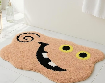 May include: A peach-colored bath mat with a cartoon face. The face has a black swirl for hair, a wide black smile with white teeth, and two yellow eyes with black pupils.