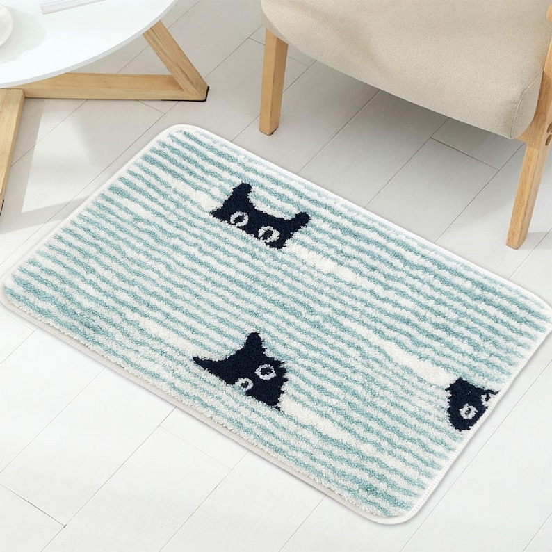 May include: A rectangular bath mat with a striped pattern in light blue and white. The mat features a repeating design of black cat faces peeking out from the stripes. The mat is placed on a white floor.