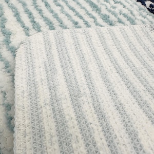 May include: Close-up of a white and blue rug with a textured surface. The rug has a pattern of diagonal stripes in shades of blue and white. The underside of the rug is visible, showing a white, patterned backing.