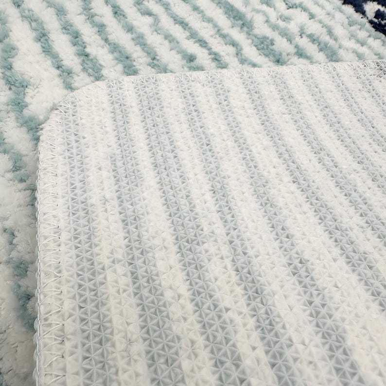 May include: Close-up of a white and blue rug with a textured surface. The rug has a pattern of diagonal stripes in shades of blue and white. The underside of the rug is visible, showing a white, patterned backing.
