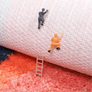 May include: A playful image showing miniature figures climbing a textured surface, possibly a rug or fabric. One figure wears a grey suit, the other an orange jumpsuit. A small ladder rests on a fluffy, coral-coloured surface, adding a whimsical touch.