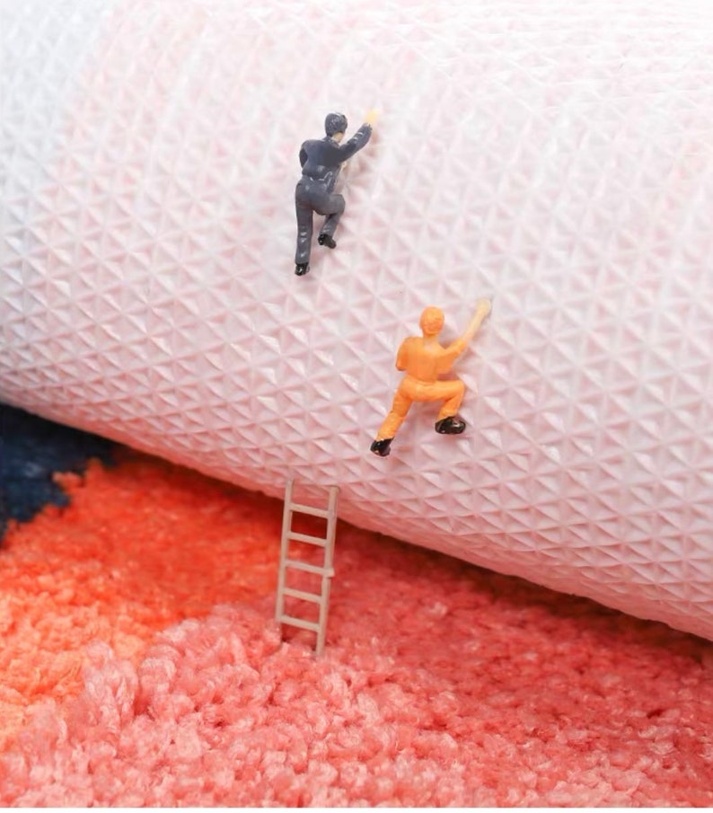 May include: A playful image showing miniature figures climbing a textured surface, possibly a rug or fabric. One figure wears a grey suit, the other an orange jumpsuit. A small ladder rests on a fluffy, coral-coloured surface, adding a whimsical touch.