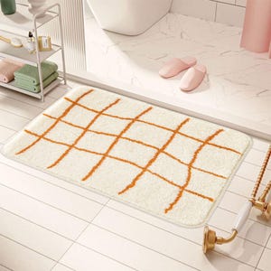Peut inclure: Tapis de bain rectangulaire crème avec un motif quadrillé orange. Le tapis est posé sur un sol carrelé blanc, suggérant un cadre de salle de bain. Le design est simple et moderne, adapté à une décoration minimaliste.