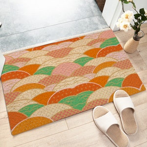 Puede incluir: Una alfombra rectangular para la puerta con un patrón geométrico colorido de formas superpuestas de color naranja, rosa, verde y blanco. El patrón se asemeja a olas o nubes. La alfombra está hecha de un material natural como el corcho o la madera.