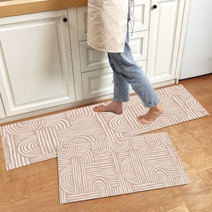 Puede incluir: Dos alfombras de cocina beige con un patrón geométrico de líneas y arcos blancos. Las alfombras se colocan delante de un armario de cocina blanco.