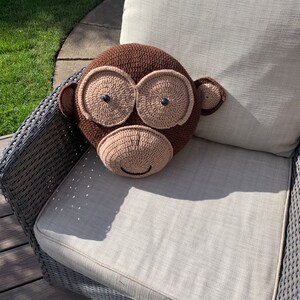 Op de afbeelding: Een bruin gehaakt aap kussen met zwarte ogen en een glimlachende mond. Het kussen ligt op een grijze rieten stoel.