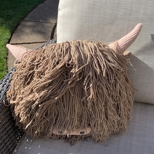 May include: A brown and pink crocheted Highland Cow pillow. The pillow is sitting on a light gray couch.