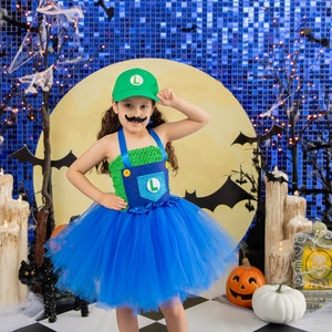 Puede incluir: Un ni&ntilde;o peque&ntilde;o vestido como un personaje de videojuego para Halloween. Lleva un tut&uacute; verde y azul, un sombrero verde con una 'L' y un bigote falso. El ni&ntilde;o est&aacute; de pie frente a un fondo azul y amarillo con murci&eacute;lagos y una luna.