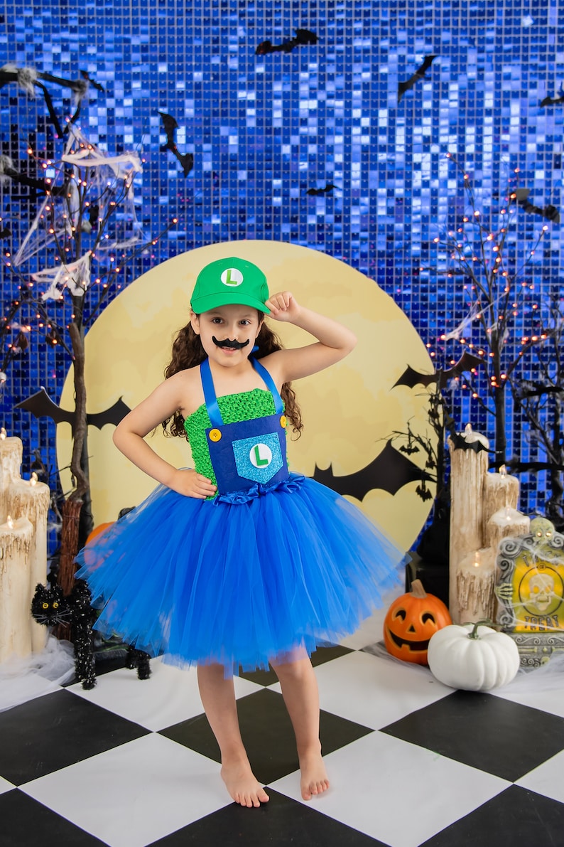 Puede incluir: Un ni&ntilde;o peque&ntilde;o vestido como un personaje de videojuego para Halloween. Lleva un tut&uacute; verde y azul, un sombrero verde con una 'L' y un bigote falso. El ni&ntilde;o est&aacute; de pie frente a un fondo azul y amarillo con murci&eacute;lagos y una luna.
