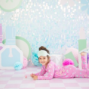 May include: A young girl wearing pink pajamas with a unicorn print and a light green sleep mask is lying on a pink and white checkered floor. She is holding a small pink bottle of nail polish. The background is a light blue and green iridescent wall with paper cutouts of nail polish bottles and a flower.