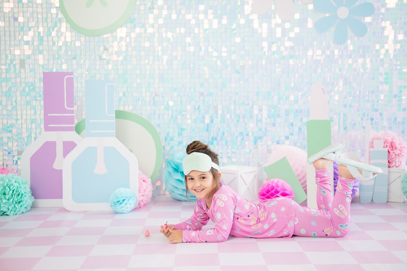 May include: A young girl wearing pink pajamas with a unicorn print and a light green sleep mask is lying on a pink and white checkered floor. She is holding a small pink bottle of nail polish. The background is a light blue and green iridescent wall with paper cutouts of nail polish bottles and a flower.
