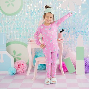 May include: A young girl wearing pink pajamas with a print of beauty products and a matching eye mask. She is standing in front of a pink vanity set with a stool and a light blue and pink background.