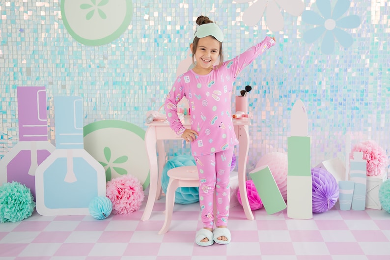 May include: A young girl wearing pink pajamas with a print of beauty products and a matching eye mask. She is standing in front of a pink vanity set with a stool and a light blue and pink background.