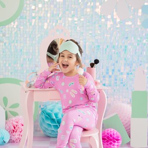 May include: A young girl wearing pink pajamas with a sleep mask on her head sits at a pink vanity table with a mirror. She is holding a makeup brush and smiling. The background is a light blue and green iridescent wall with decorative paper flowers.