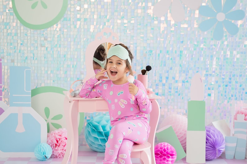 May include: A young girl wearing pink pajamas with a sleep mask on her head sits at a pink vanity table with a mirror. She is holding a makeup brush and smiling. The background is a light blue and green iridescent wall with decorative paper flowers.